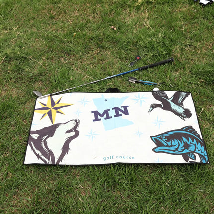 Eliteshine MH-B golf towel laid flat on grass showing full rectangular custom print — multiple design examples including golf course and nature themes