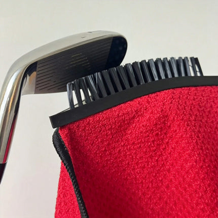 Close-up of Eliteshine MH-E golf towel showing built-in brush scrubbing a golf iron club face — red waffle cotton towel with black overlock edge and rubber bristle brush